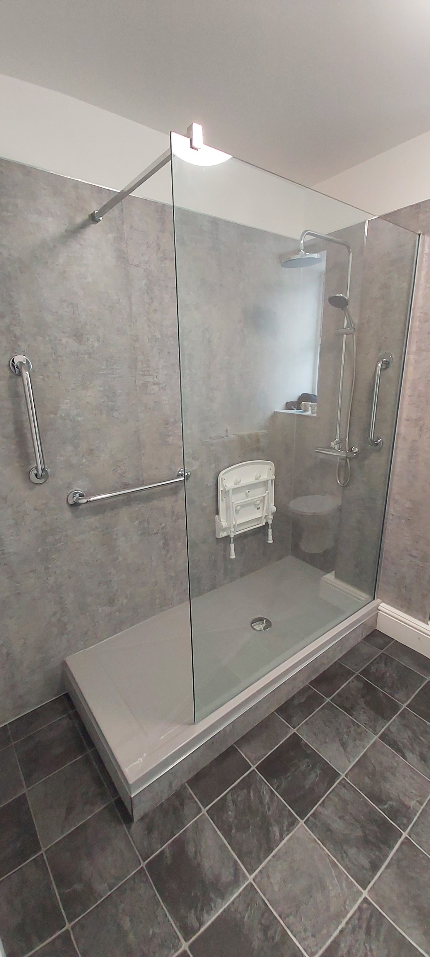 The finished walk-in shower with grey wall panels, a glass screen, grab rails and a fold-down seat